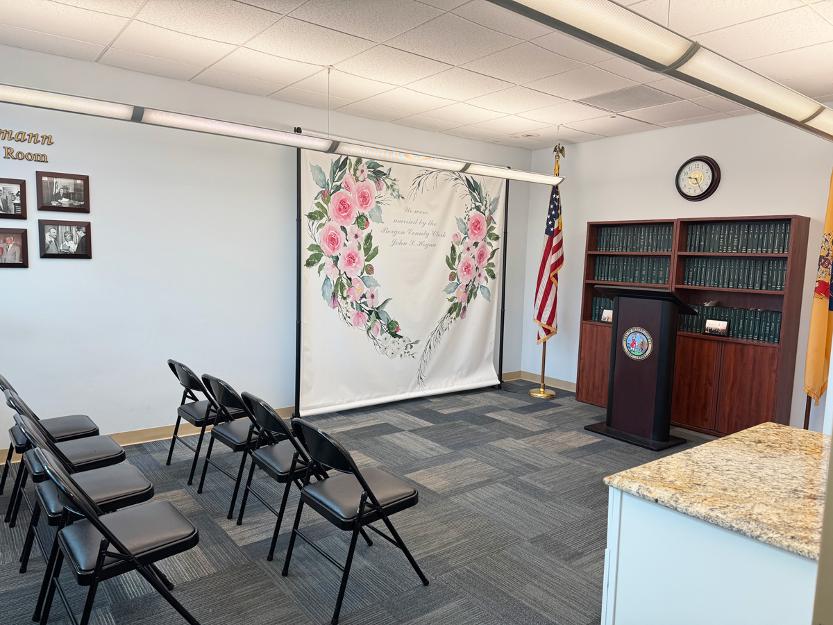 Ceremony room at the Bergen County Clerk's Office, with guest chairs facing a floral backdrop, podium, and American flag.